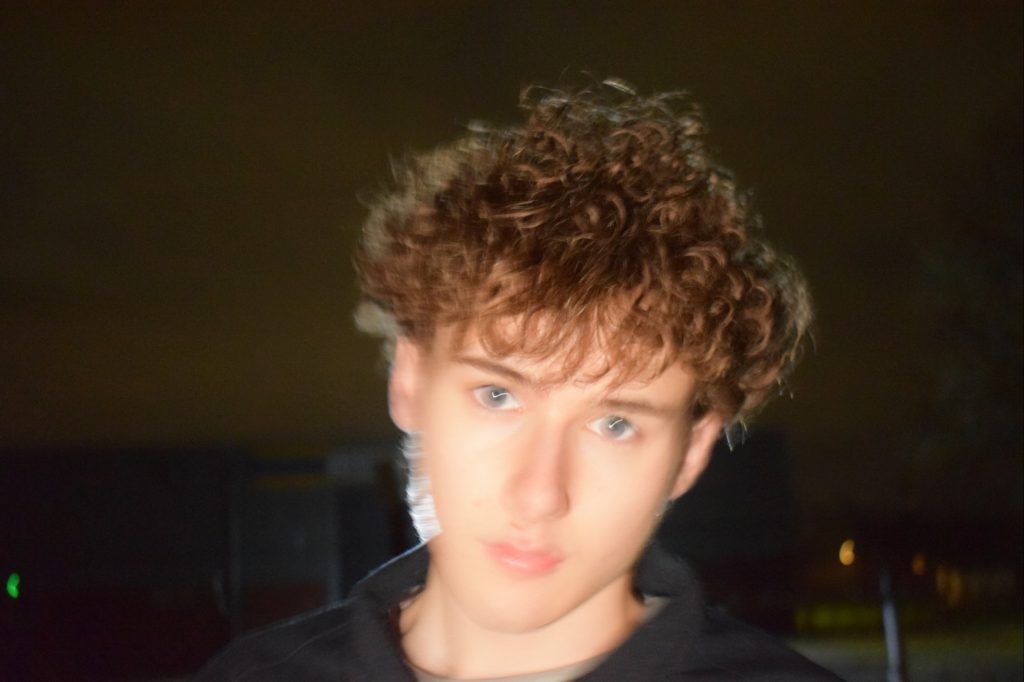 Closeup shot of Elliott Guy, musician, in January 2024. At night / nighttime, face, head slightly tilted, flash camera brown sky. black jumper hoodie. streetlight and building blurred but visible in background